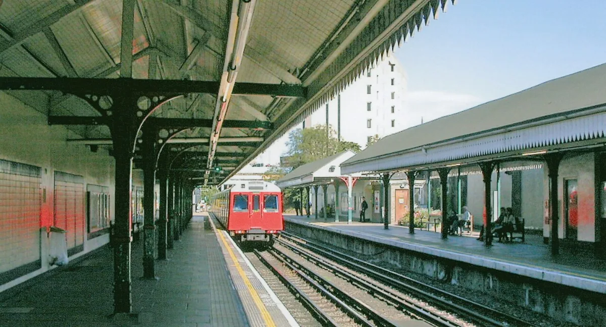West Kensington Station, with eastbound train. View eastward, towards Earls Court, Central and East London, also Edgware Road via High Street Kensington: LUL District Lines.